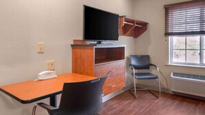 A room with a wooden desk, a chair, a television mounted on a wooden cabinet, a telephone, and a window with blinds. - Studio 6 Extended Stay Wichita South KS (Wichita)
