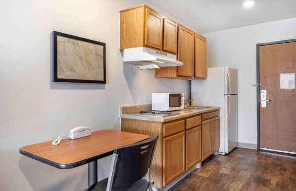 A compact kitchenette with wooden cabinets, a microwave, a refrigerator, a small table with a chair, and a phone on the table.