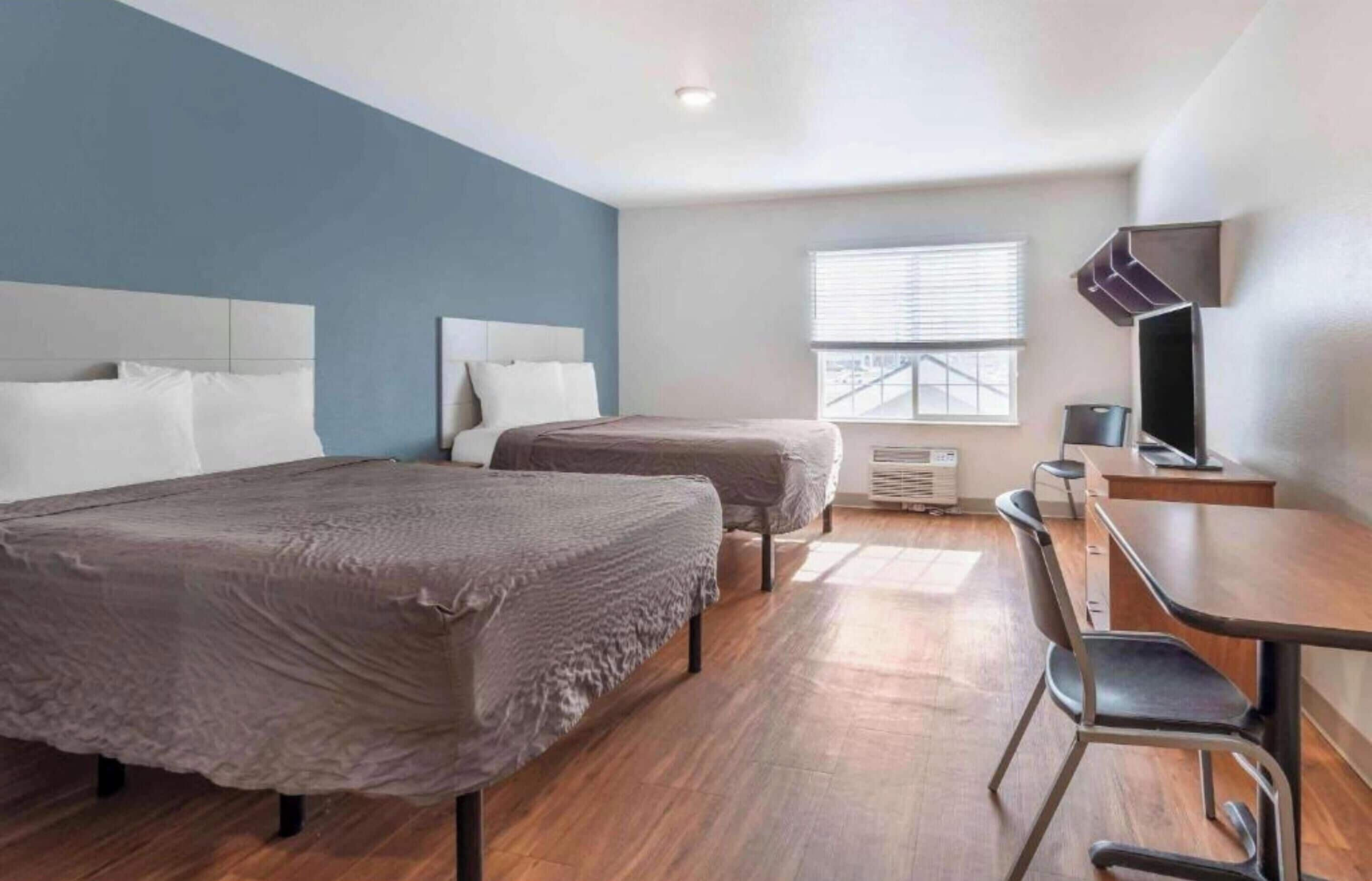 A hotel room with two beds, a desk, a chair, a TV, and a window with blinds.