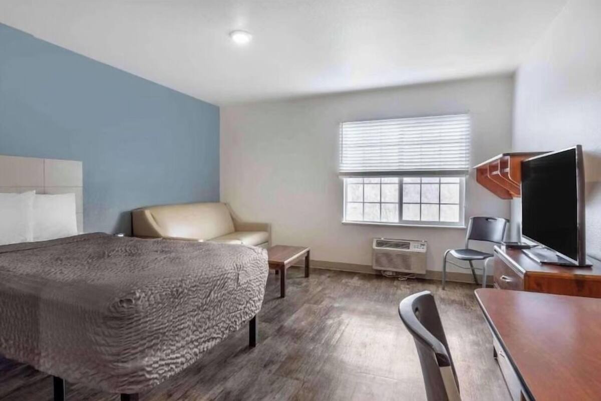Une chambre d’hôtel équipée d’un lit, d’une chaise, d’un bureau, d’une télévision et d’une fenêtre avec des stores.