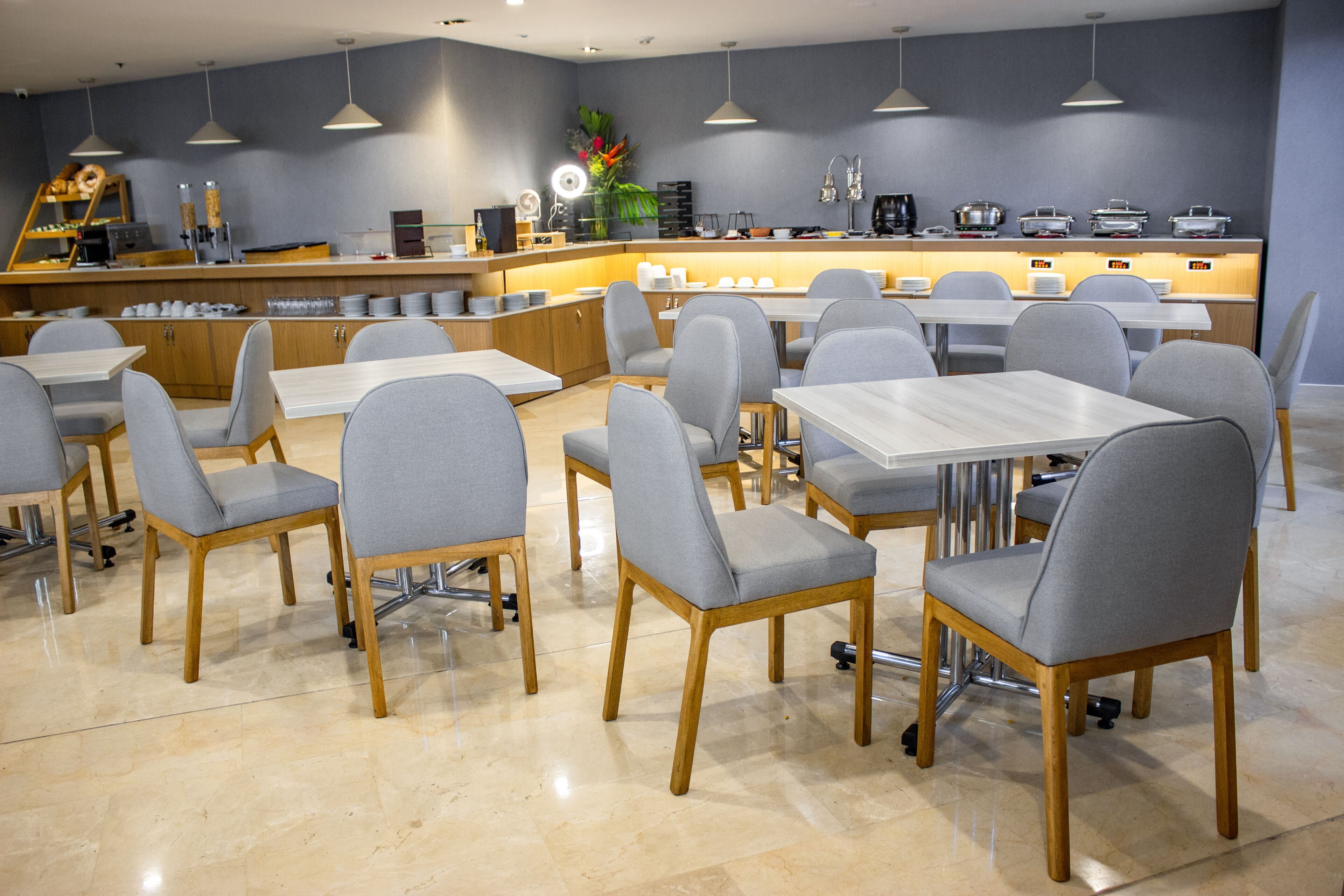 A modern dining area with grey upholstered chairs and wooden tables, a counter with various appliances, and a decorative plant.