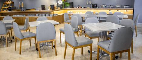 A modern dining area with grey upholstered chairs and wooden tables, a counter with various appliances, and a decorative plant.