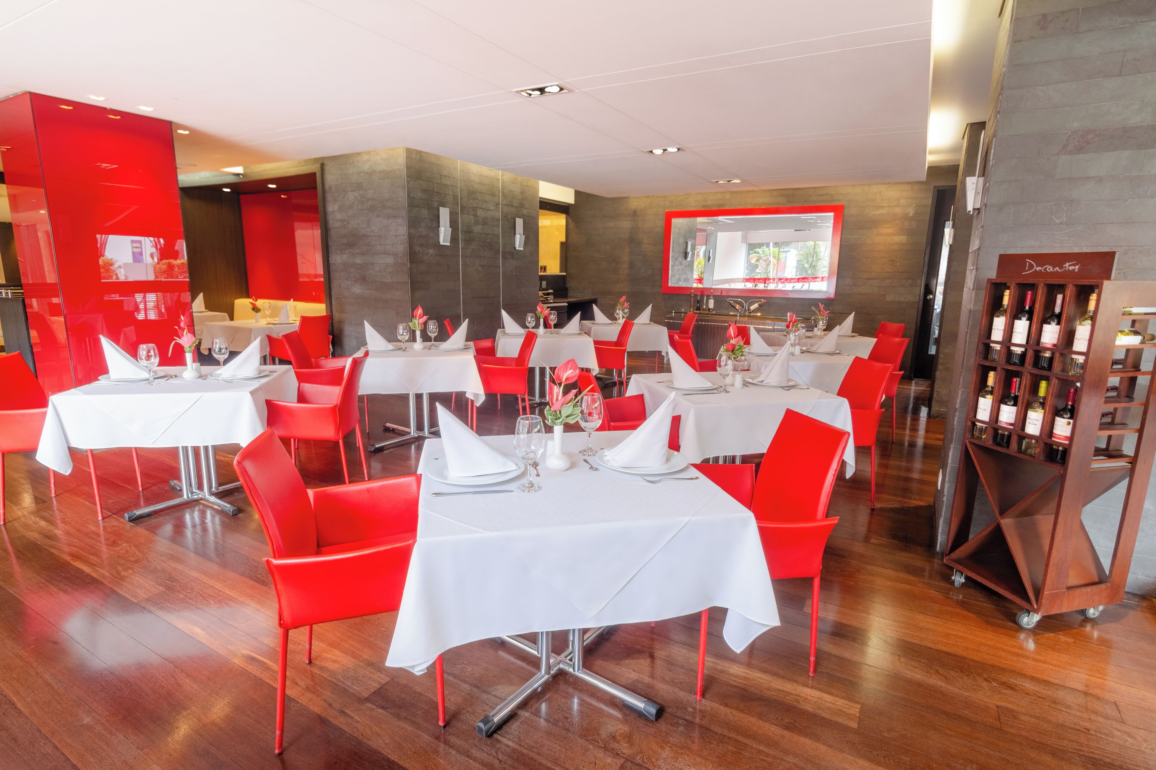 Ein Restaurant mit mehreren gedeckten Tischen, roten Stühlen und einer Weinauslage.