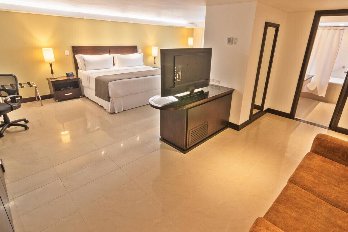 A hotel room with a bed, a desk with a television, a chair, and a sofa.