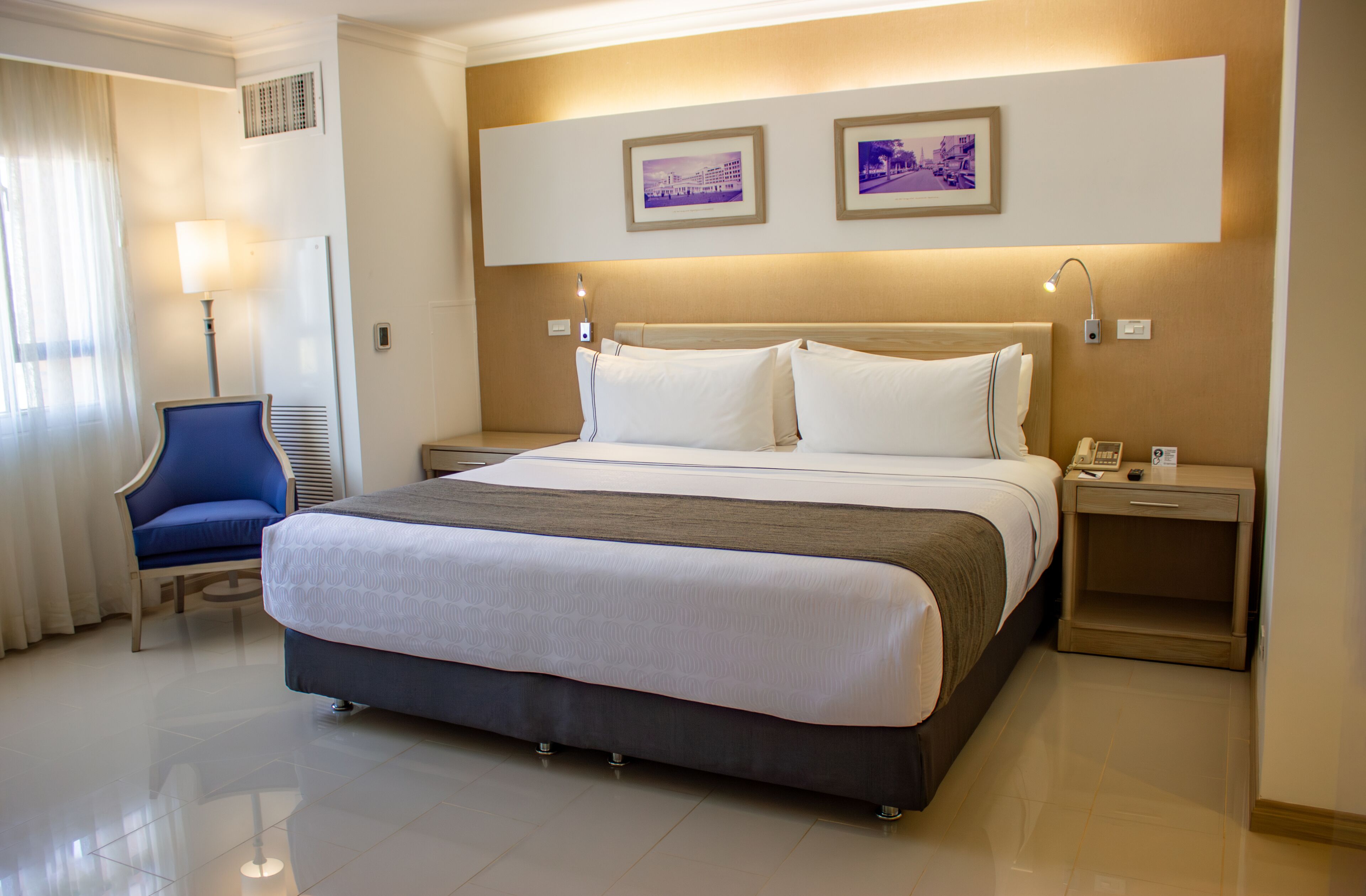 Habitación de hotel con una cama grande, una mesita de noche con lámpara, una silla y dos cuadros en la pared.
