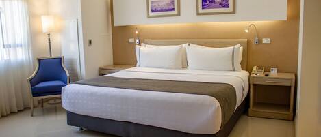 A hotel room with a large bed, a bedside table with a lamp, a chair, and two framed pictures on the wall.