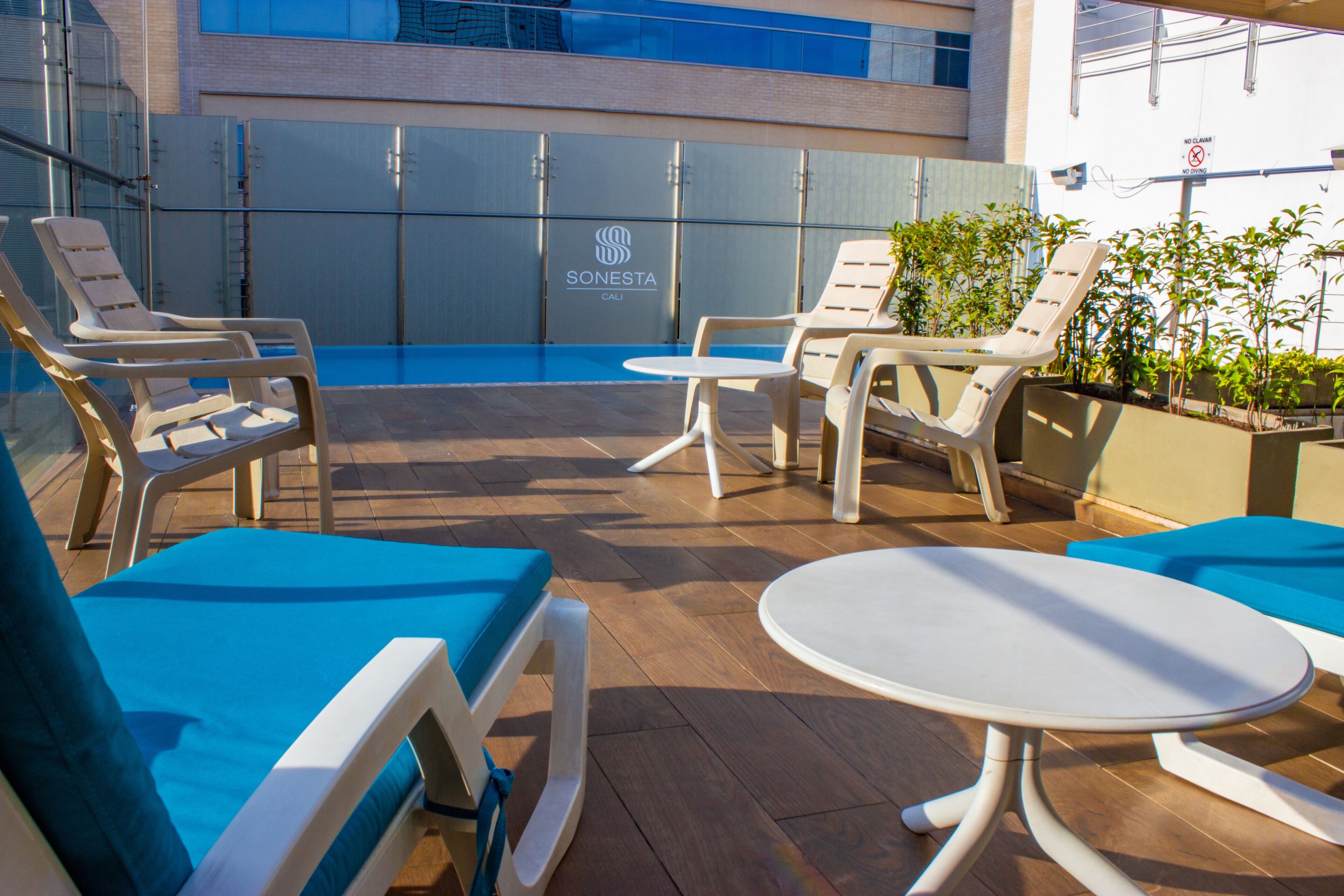 A rooftop pool area with lounge chairs and a small table, a glass wall with a logo, and a building in the background.