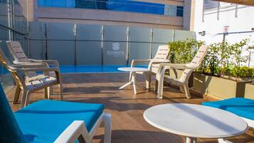 A rooftop pool area with lounge chairs and a small table, a glass wall with a logo, and a building in the background.