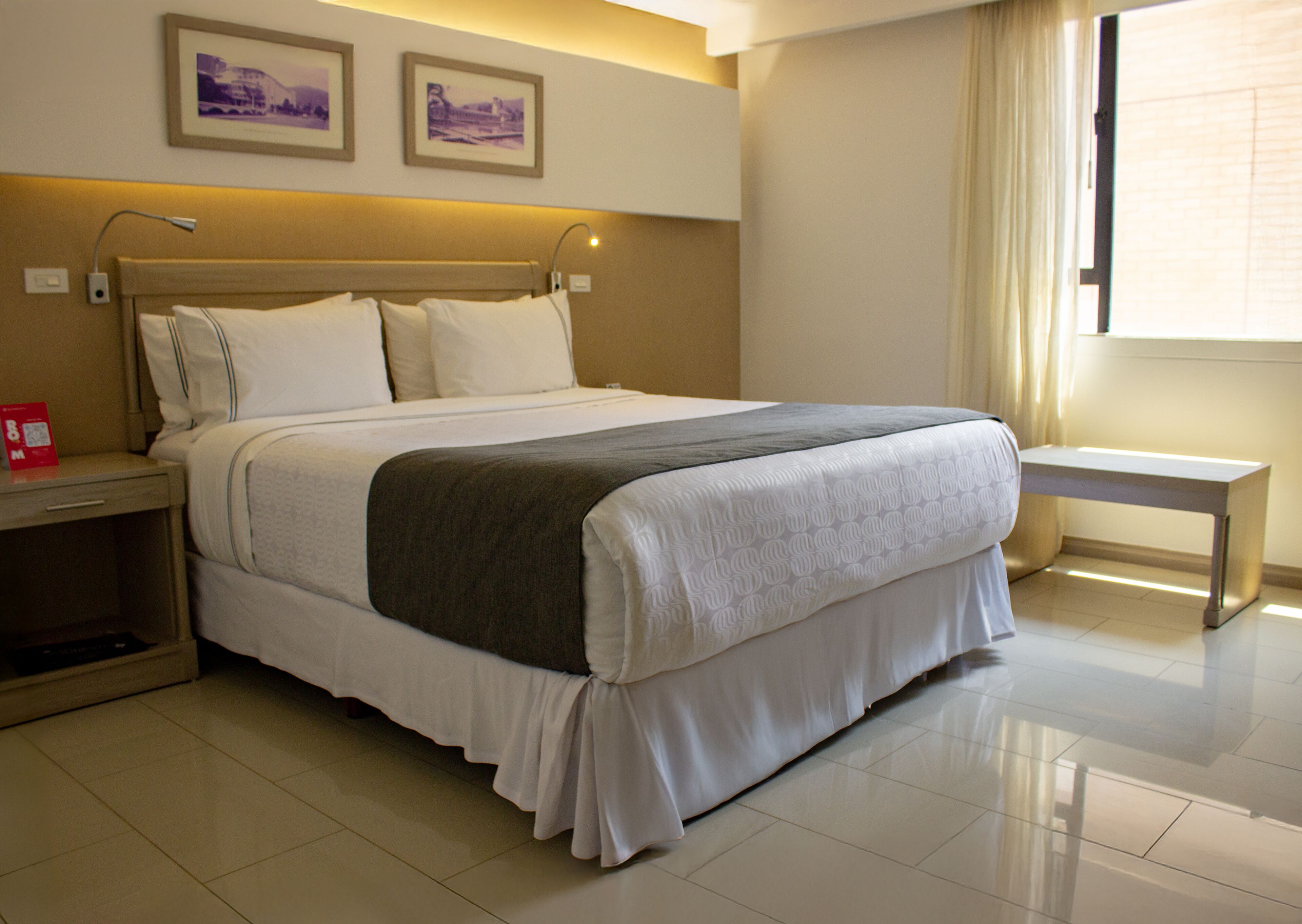 A neatly made bed with a grey blanket, a bedside table with a lamp, and a small bedside table with a red object.