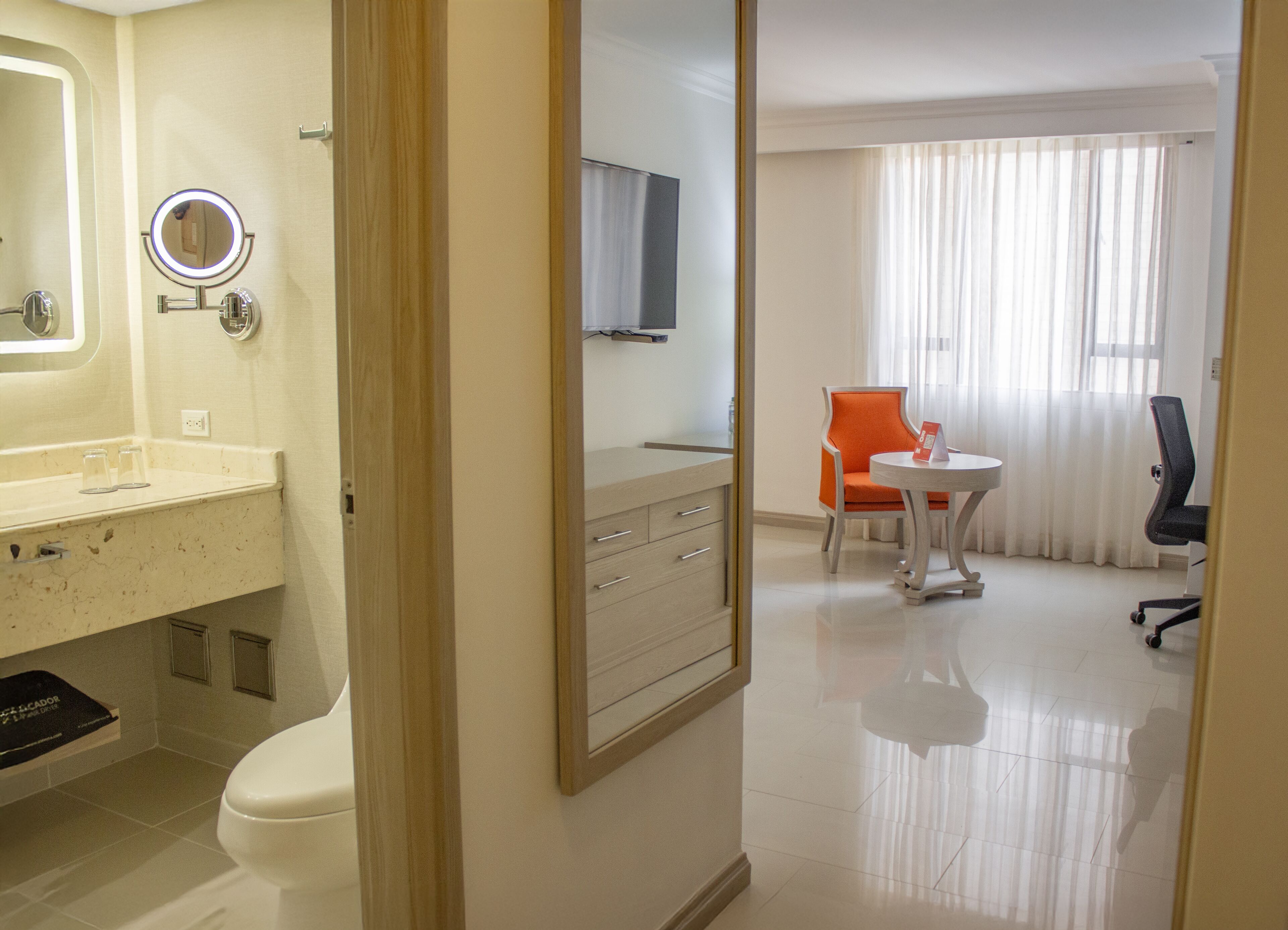A hotel room with a bathroom, a small table with a chair, and a large window with curtains.
