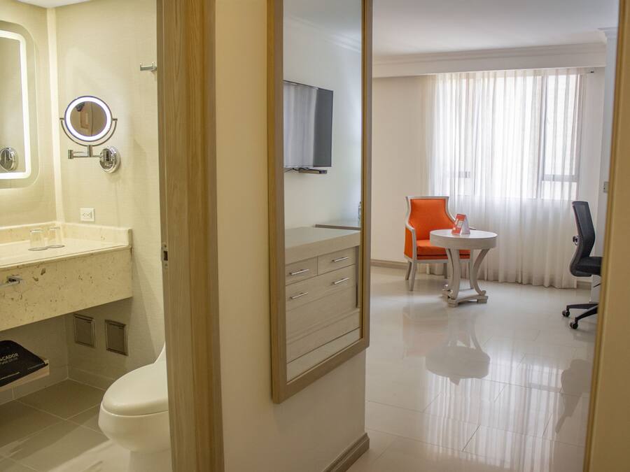A hotel room with a bathroom, a small table with a chair, and a large window with curtains.