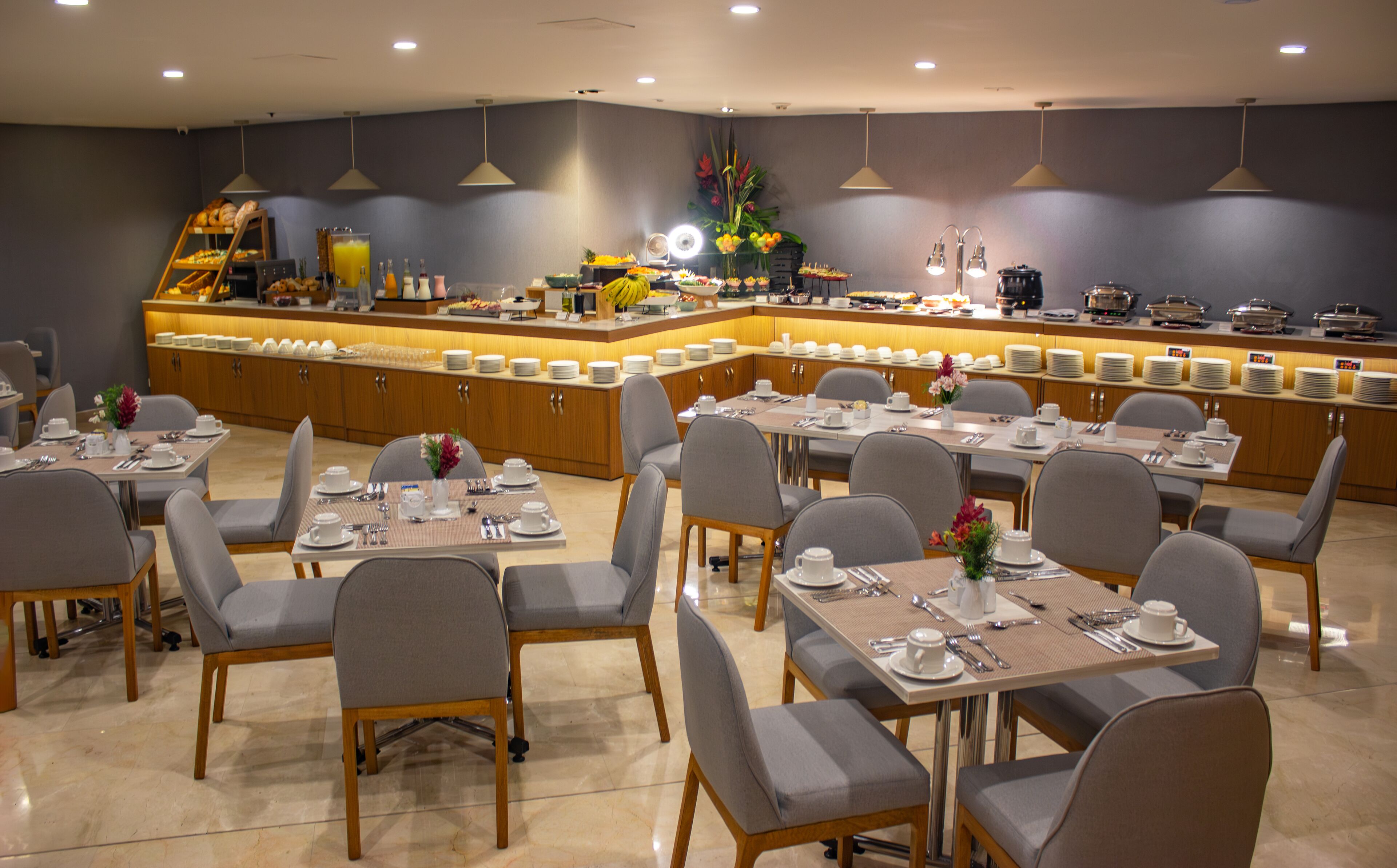 Uma sala de jantar de hotel com várias mesas e cadeiras, um balcão de buffet e diversas opções de comida e bebida.