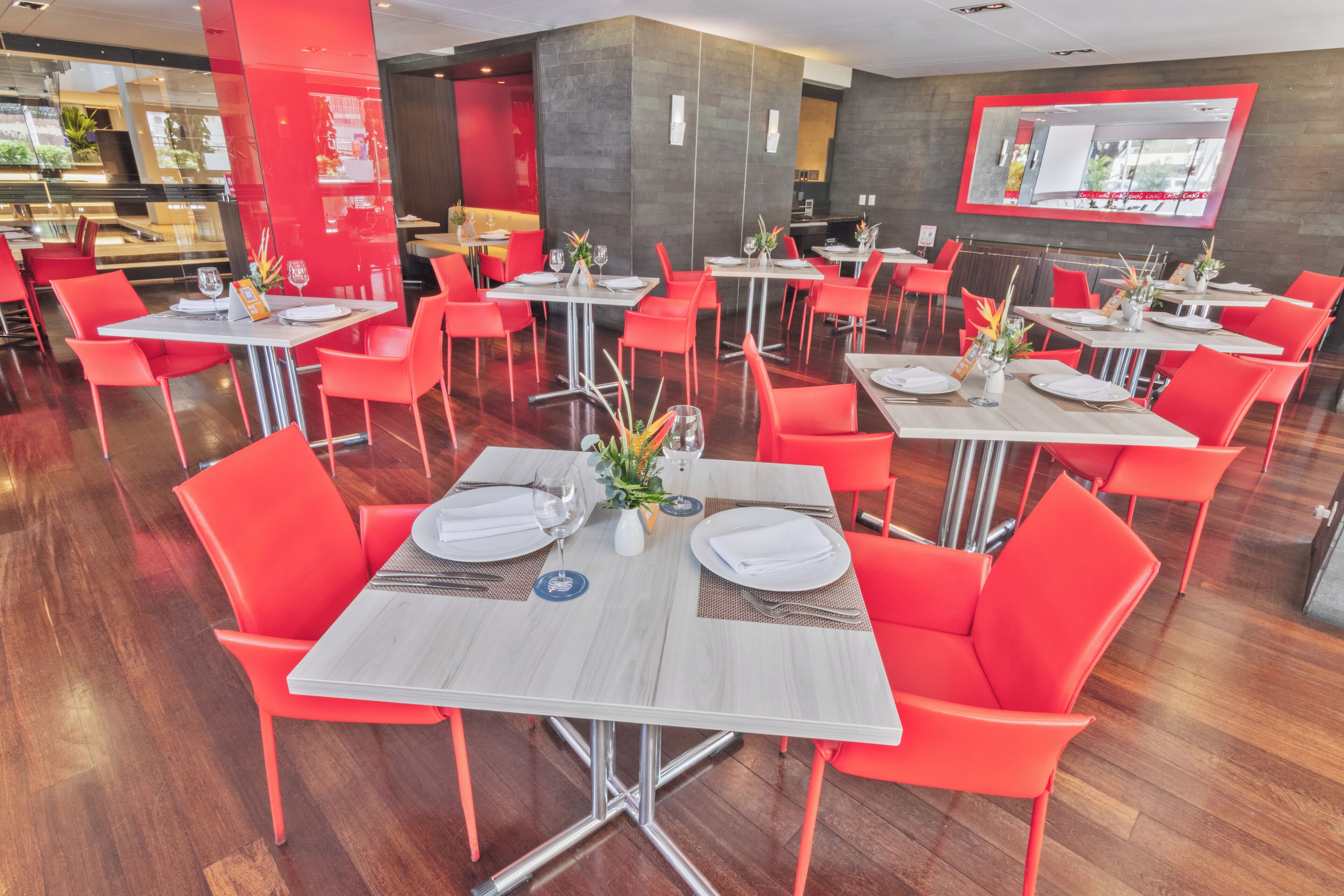 Een modern restaurant met rode stoelen en witte tafels, met een glanzende houten vloer en een grote spiegel aan de muur.
