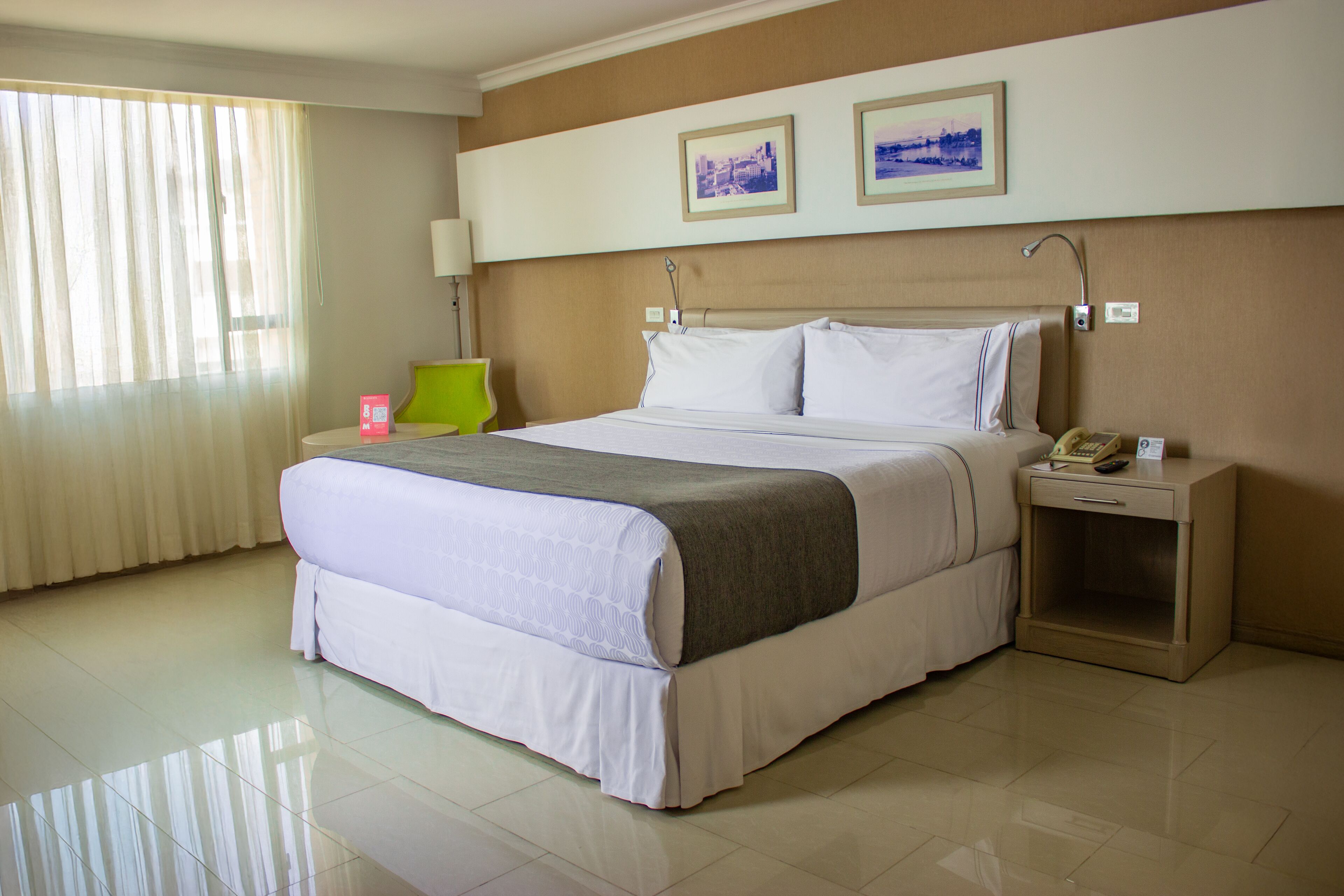 Una habitación de hotel con una cama grande, una mesita de noche con lámpara, un buró con teléfono y dos cuadros en la pared.