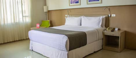 Una habitación de hotel con una cama grande, una mesita de noche con lámpara, un buró con teléfono y dos cuadros en la pared.