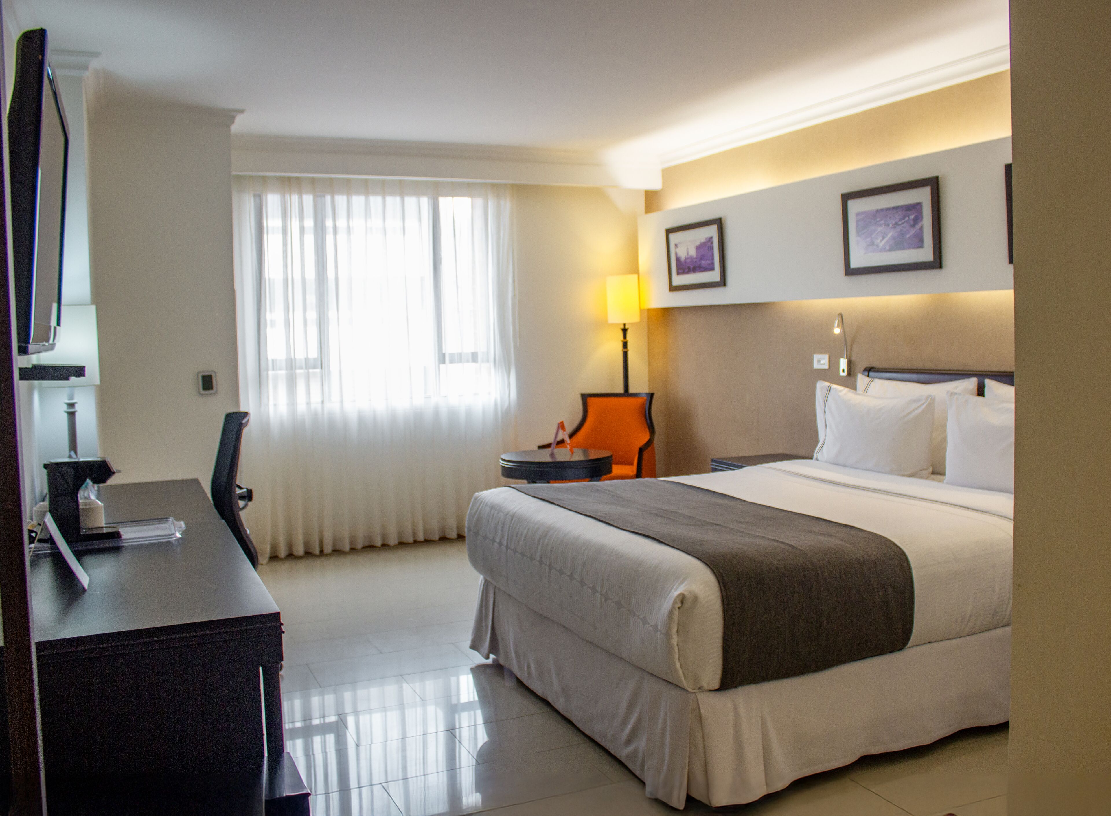 Habitación de hotel con cama, escritorio, silla, lámpara y dos cuadros en la pared.