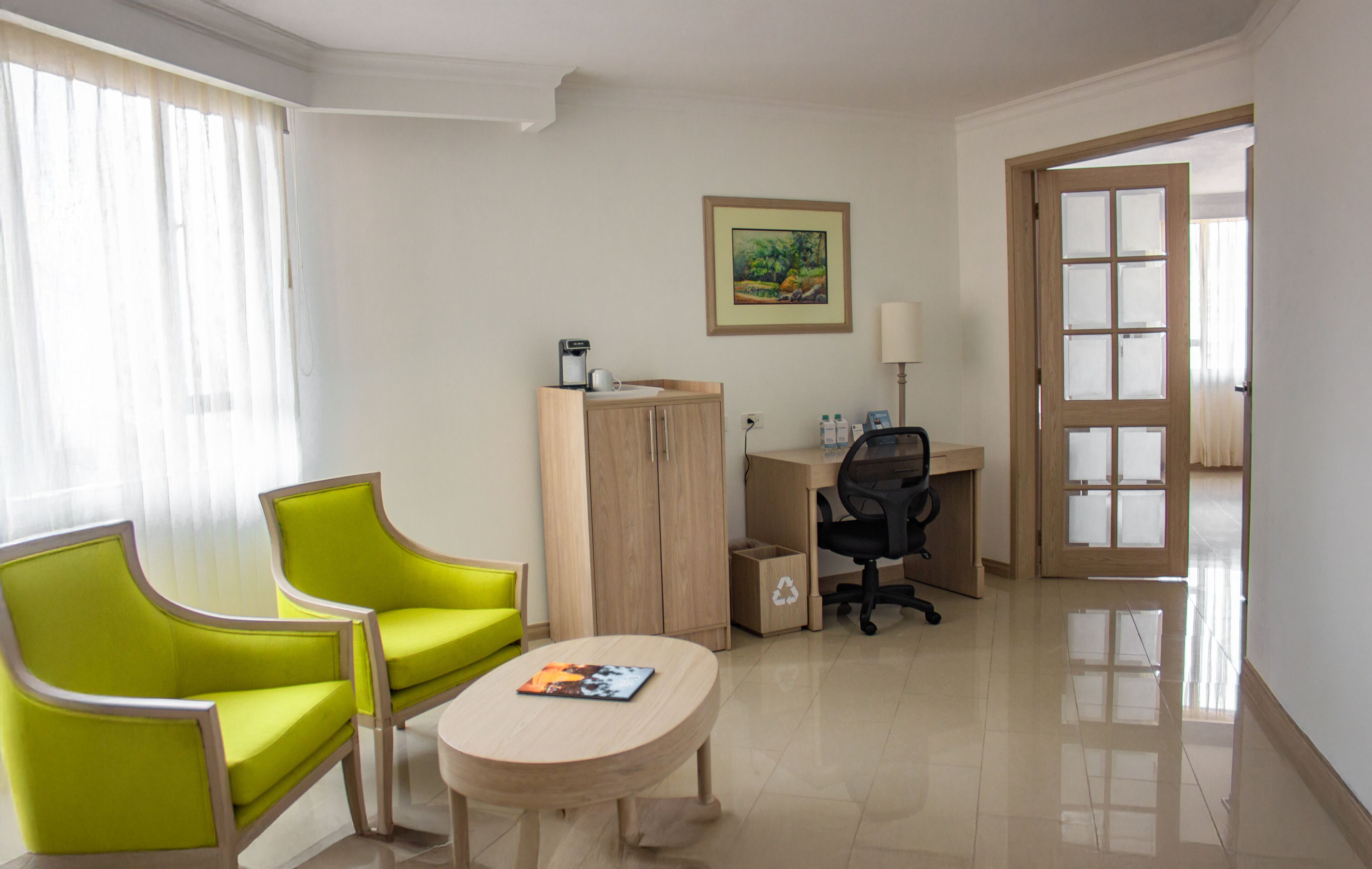 A hotel room with a desk, two green chairs, a small round table, a cabinet, and a painting on the wall.