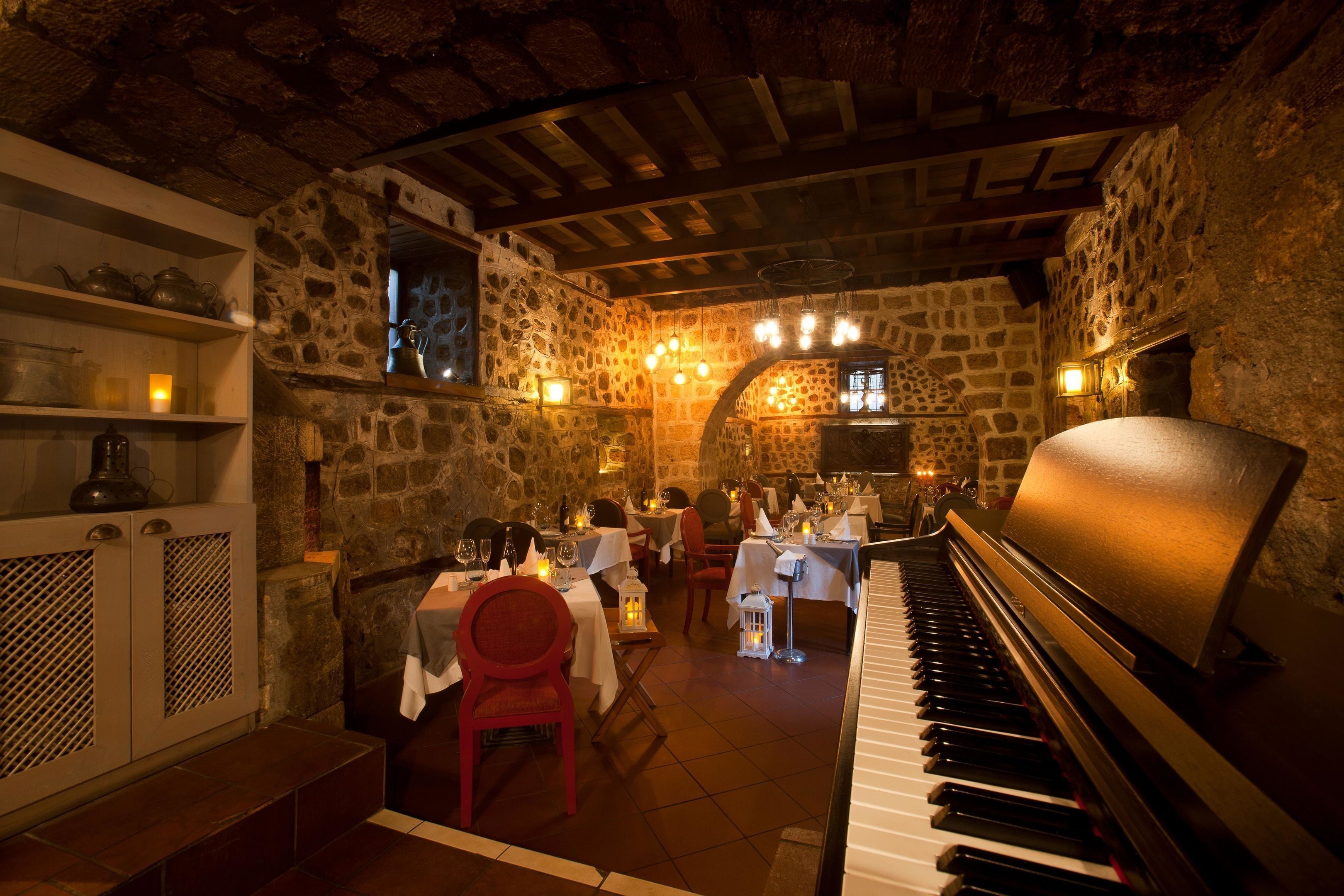 A cozy restaurant with stone walls, wooden beams, and a piano. Tables are set with candles and dinnerware.