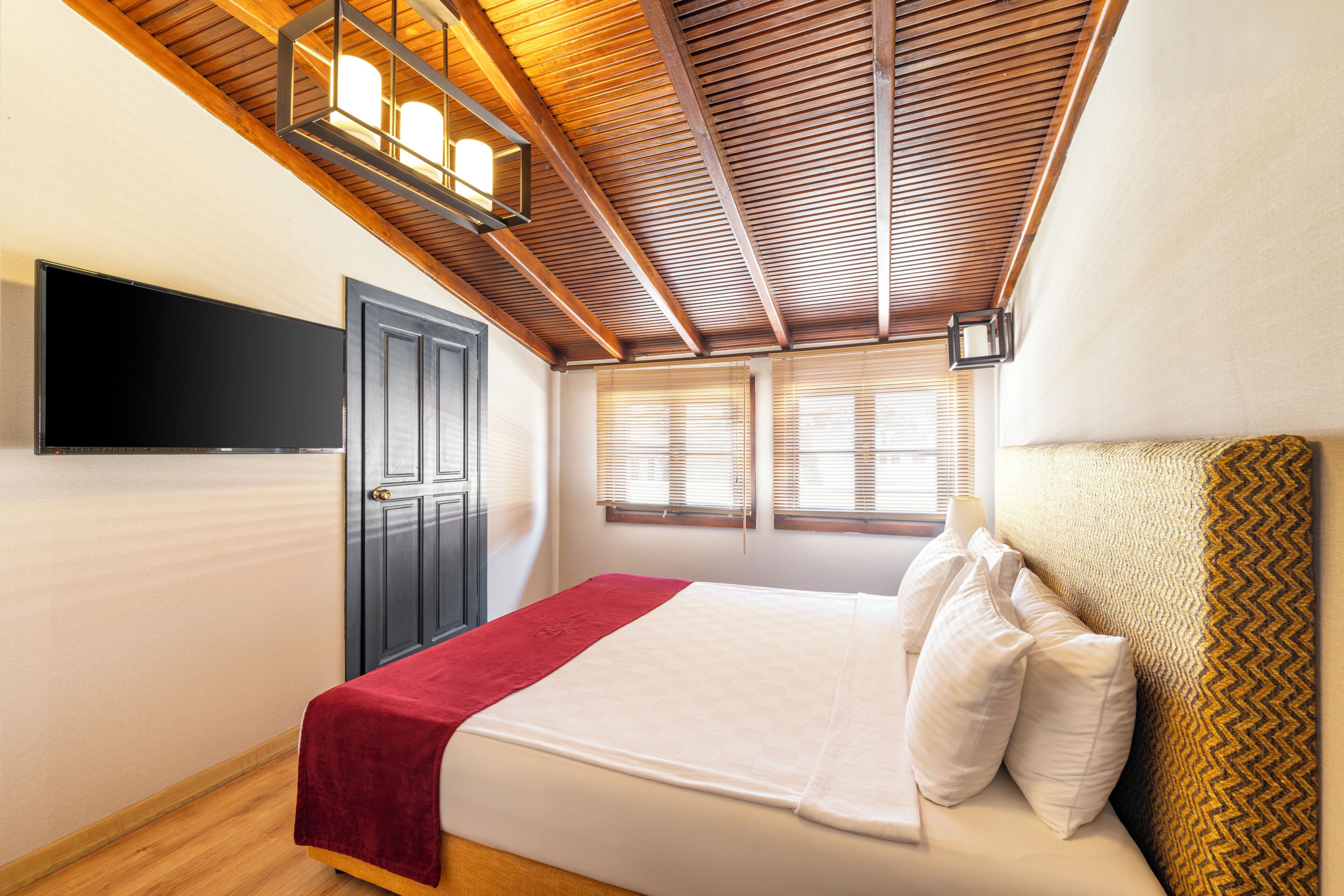 A bedroom with a bed, a television mounted on the wall, a door, and windows with blinds.