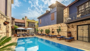 Una piscina con sillas de descanso y sombrillas, rodeada de edificios con fachadas de piedra y balcones.