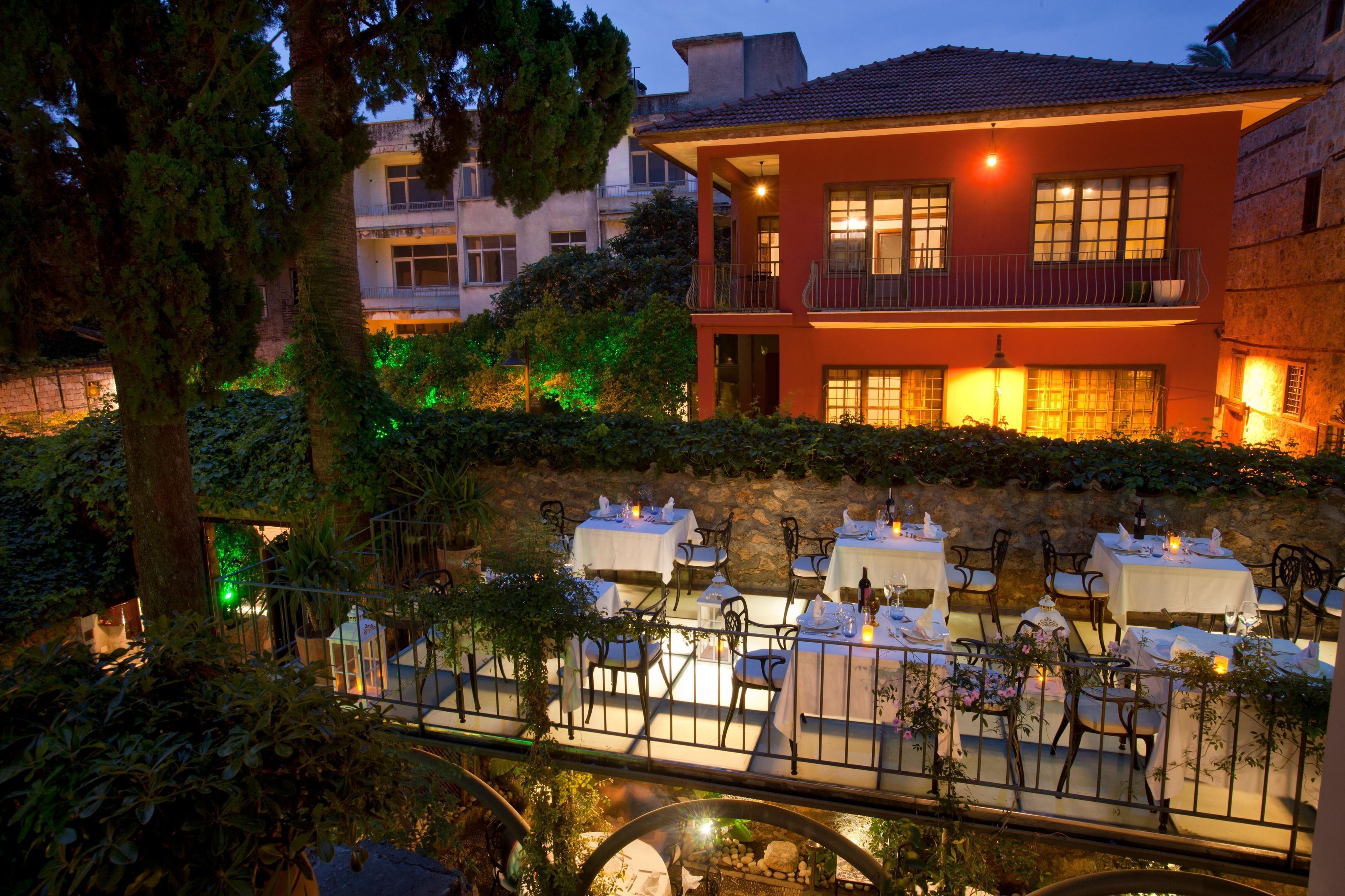 An outdoor dining area with tables and chairs, surrounded by greenery and a stone wall.