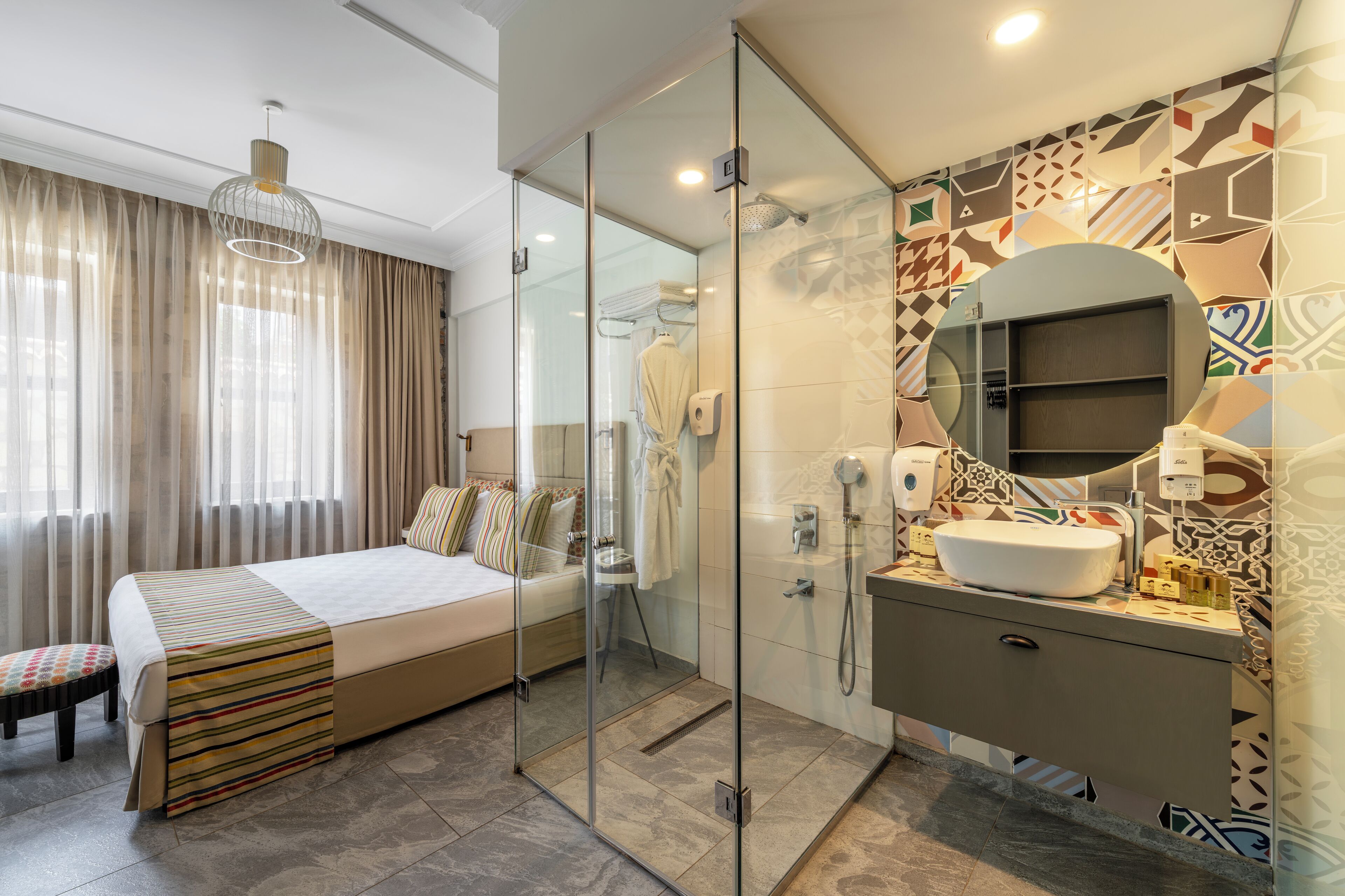 Una habitación de hotel moderna con una cama grande, cabina de ducha con paredes de vidrio, tocador con espejo y azulejos decorativos en las paredes.