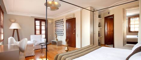 A hotel room with a four-poster bed, a sitting area with a chair and a small table, a lamp, and a decorative wall piece.