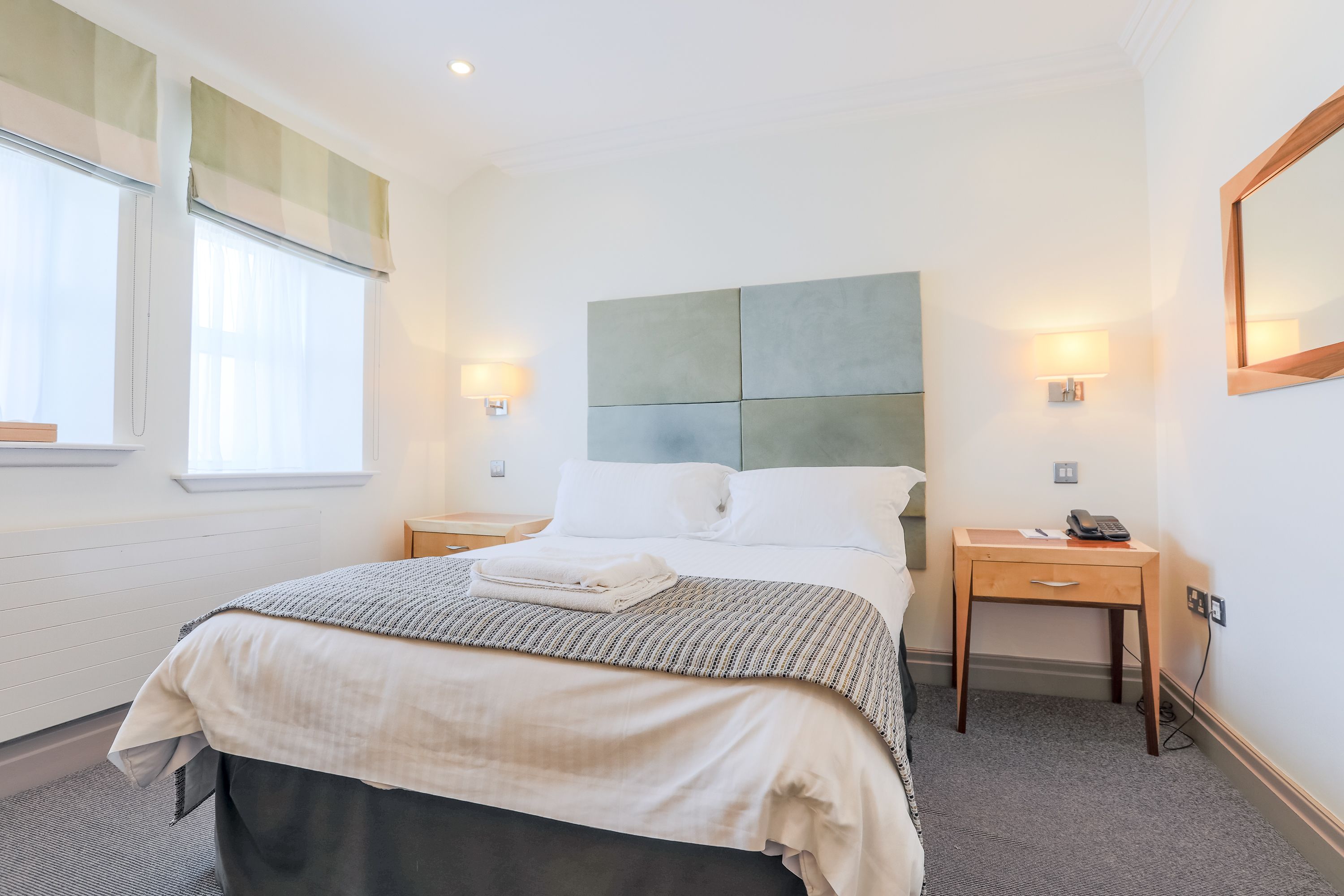 A neatly made bed with a grey and white patterned bedspread, two bedside tables with lamps, and a wooden nightstand with a phone.