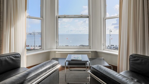 A room with two black leather sofas, a small table, and a view of the sea through large windows. - The Claremont  (Douglas)