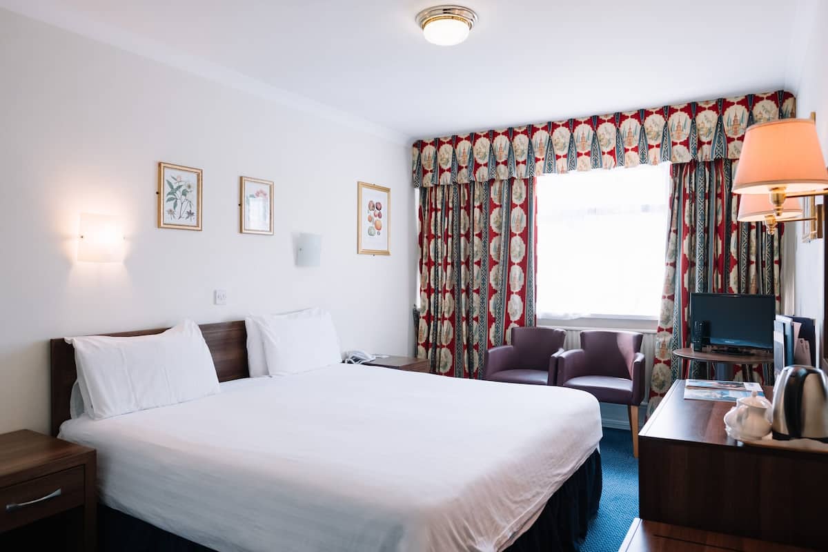 A hotel room with a large bed, a desk with a lamp, a chair, and a television.