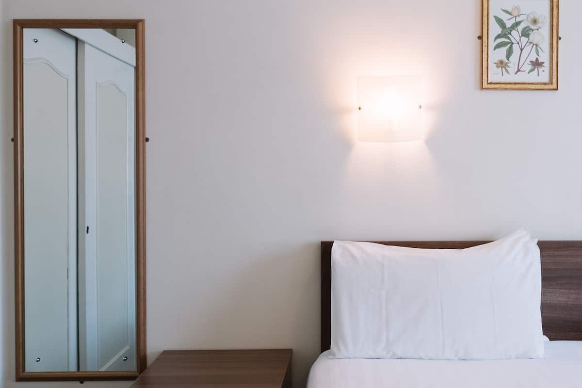 A bedroom with a wooden nightstand, a bedside table, a bed with white bedding, a wall-mounted light, and a framed botanical illustration.