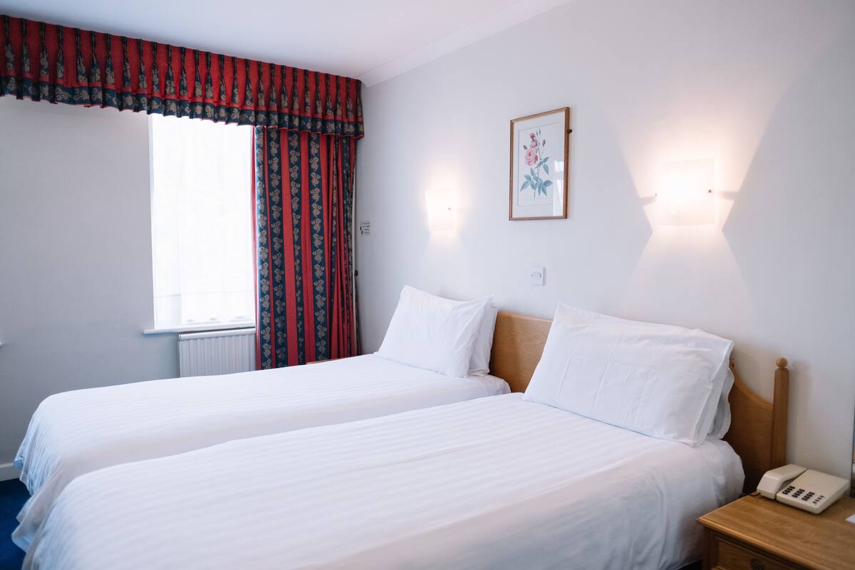 A hotel room with two single beds, a wooden headboard, a bedside telephone, and a window with red curtains.