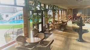 A covered outdoor seating area with wicker furniture, large windows, and potted plants. - Apollo Jersey (St. Helier)