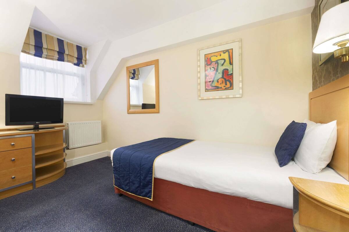 A hotel room with a bed, a desk with a TV, a mirror, and artwork on the wall.