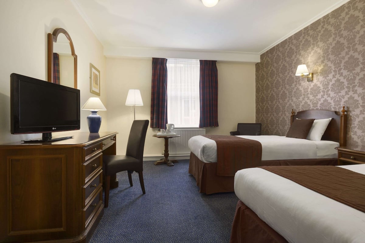A hotel room with a television, a desk with a lamp, two beds, a chair, and a window with curtains.