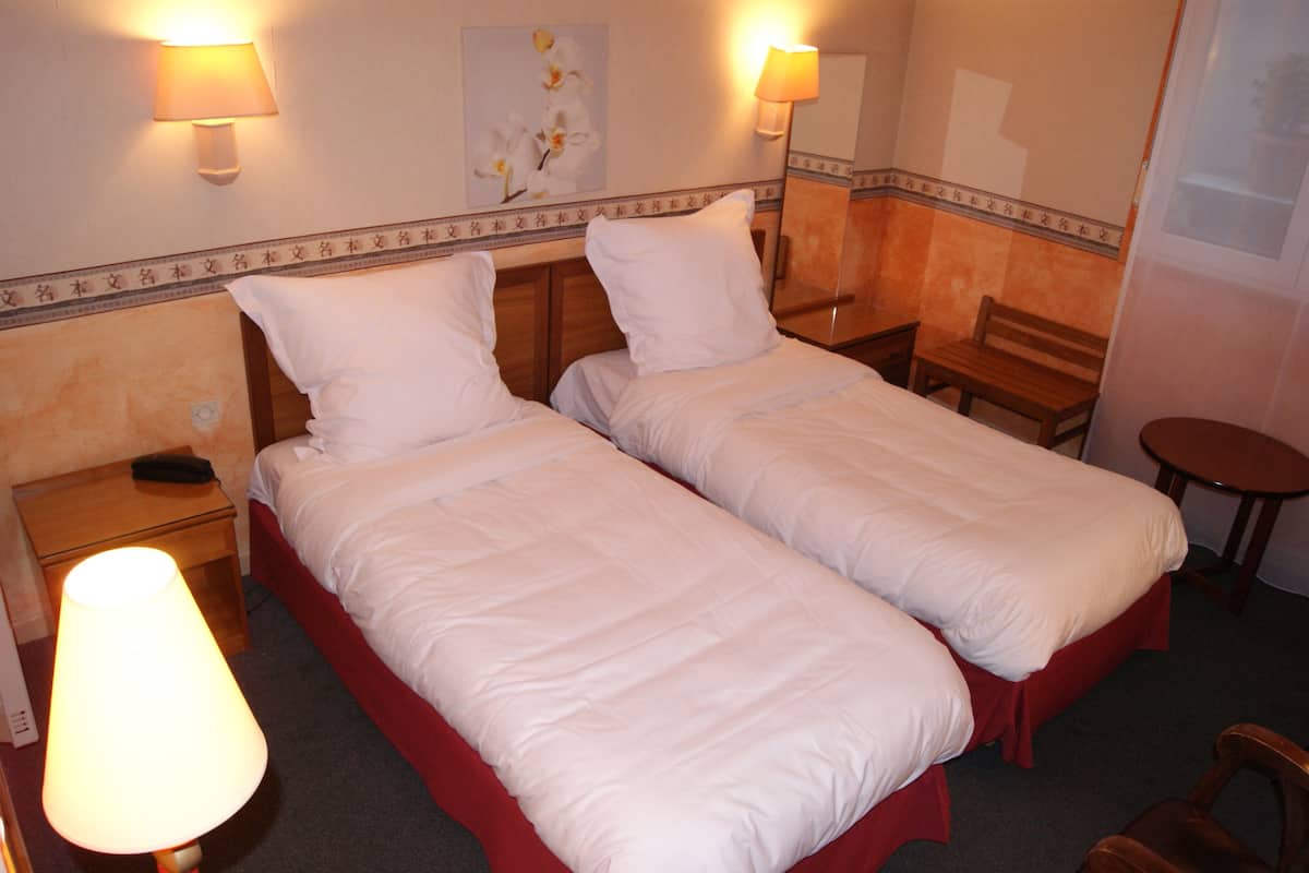 Une chambre d’hôtel avec deux lits simples, une table de chevet avec une lampe, une chaise et une petite table.