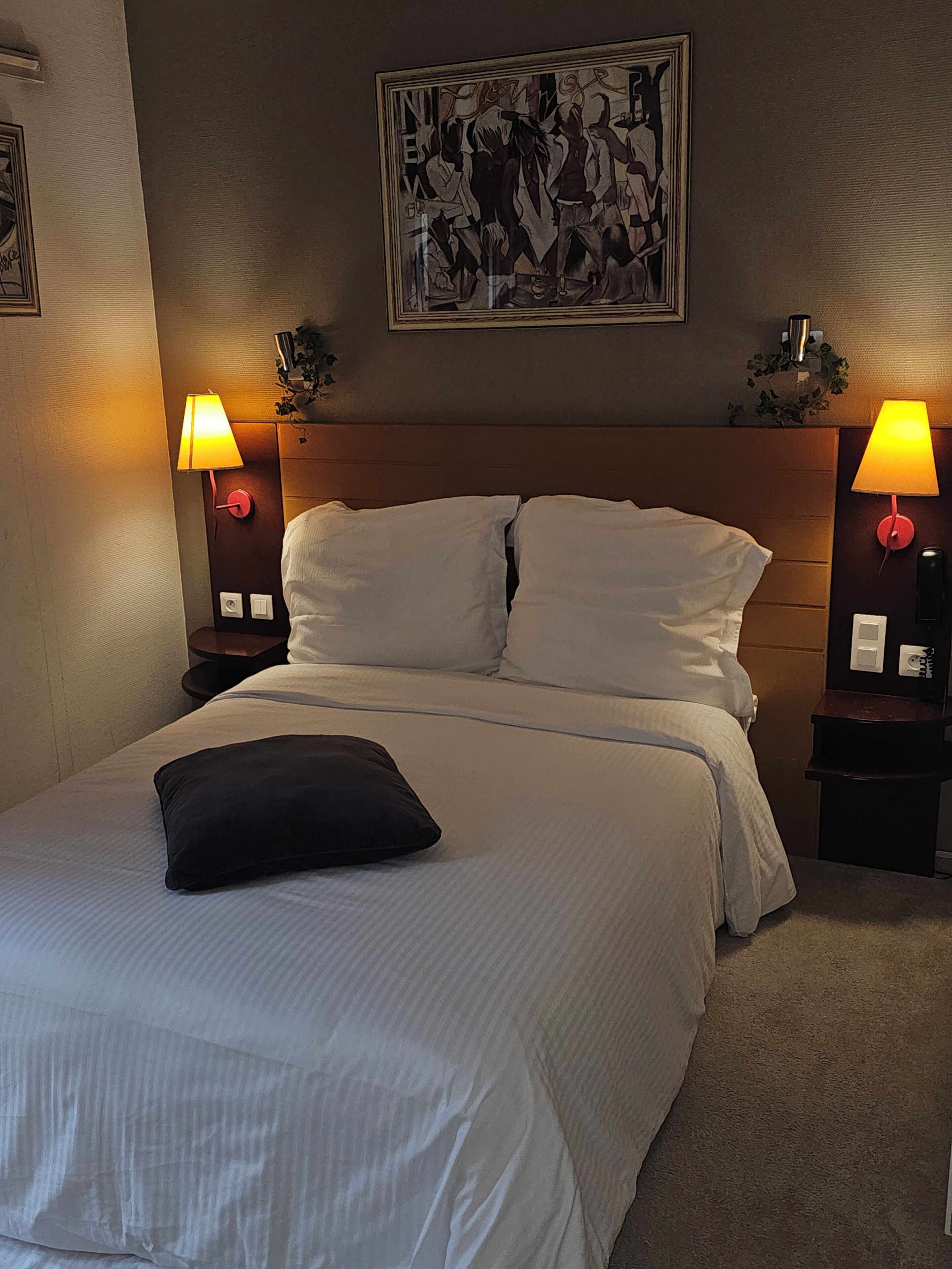 A neatly made bed with white linens and a black pillow, flanked by two bedside tables with lamps, a framed artwork above the bed, and a wall-mounted light fixture.