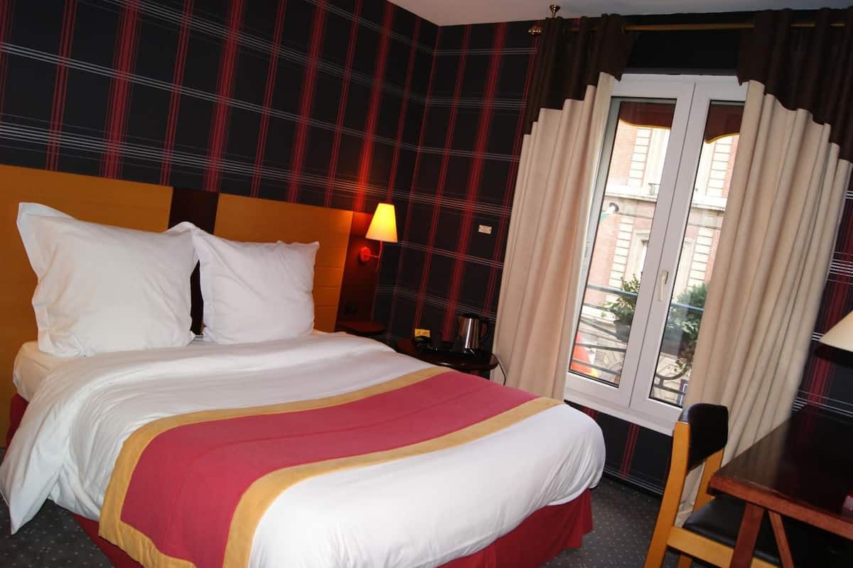 Une chambre d’hôtel avec un lit, une table de chevet, une lampe, une chaise et une fenêtre avec des rideaux.