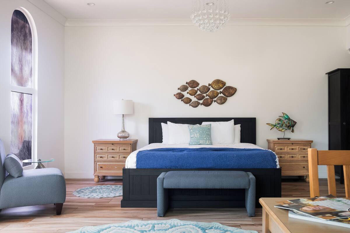 A bedroom with a large bed, two bedside tables, a chair, a bench, a chandelier, and a wall art piece.