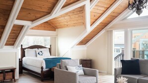Dormitorio tipo loft con techo inclinado, cama con manta azul, sillón gris, mesa de centro de madera y vistas a edificios.