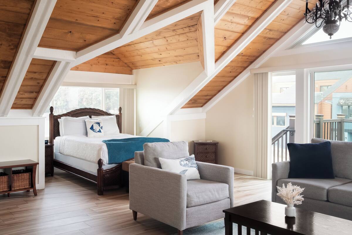 A loft bedroom with a sloped ceiling, a bed with a blue throw blanket, a grey armchair, a wooden coffee table, and a view of buildings outside.