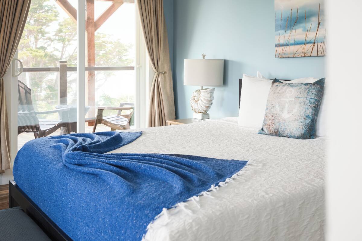 A bedroom with a bed, a nightstand, a lamp, a painting, and a view of the outdoors.