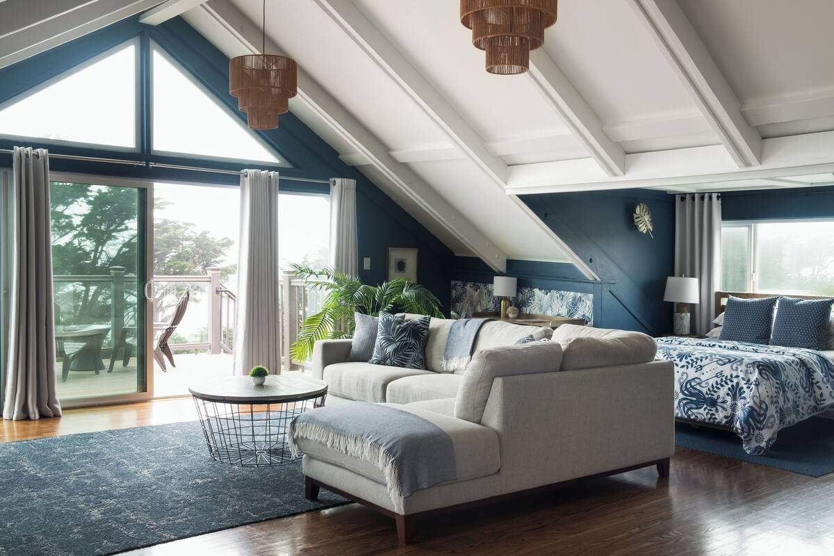 A modern living room with a sofa, a coffee table, and a bed with a patterned bedspread. There is a large window with curtains, a plant, and a hanging light fixture.