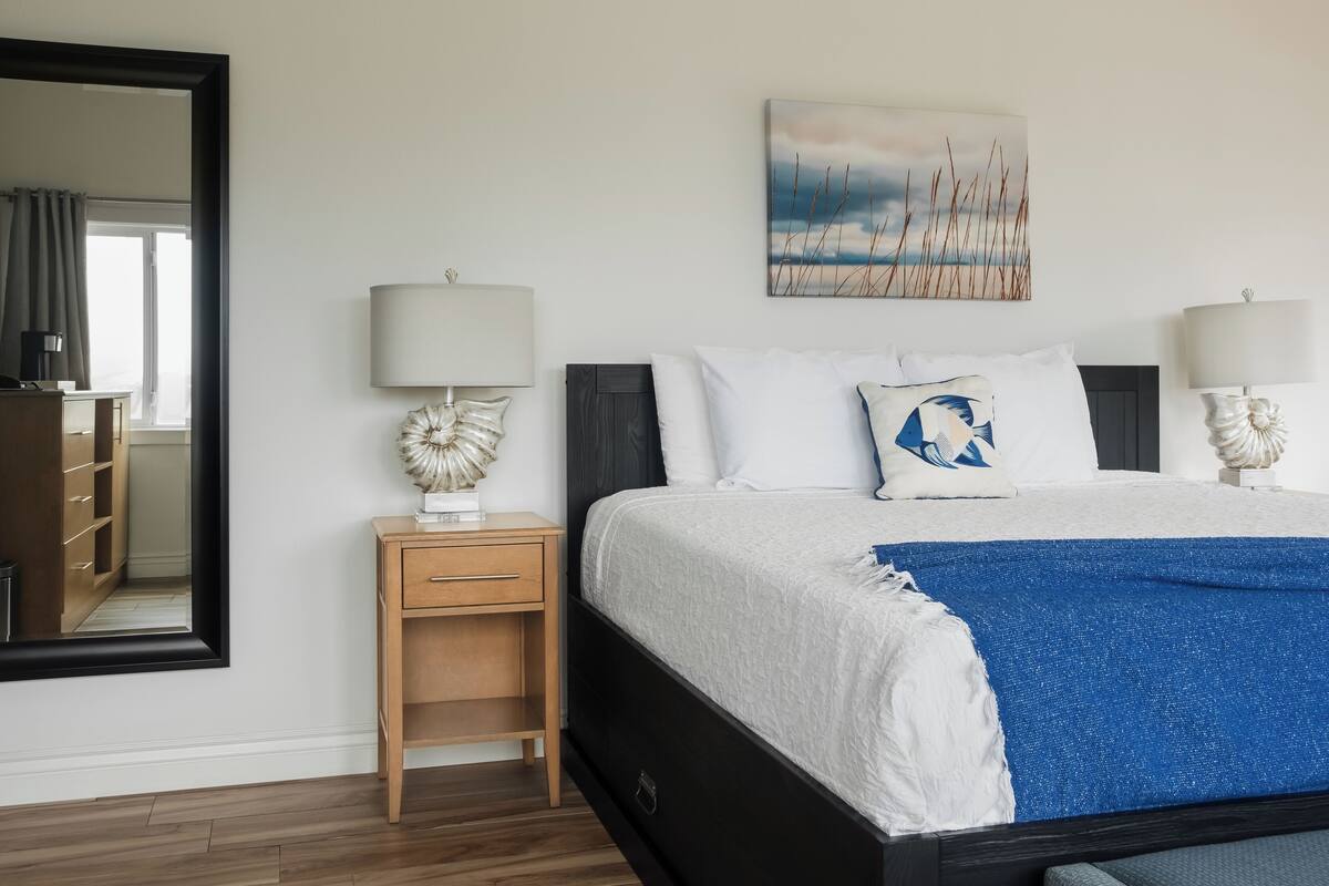 A neatly arranged bedroom with a large bed, bedside lamps, a small nightstand, a mirror, and a wall-mounted painting.