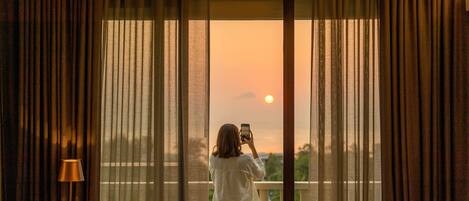 Una habitación con un ventanal grande, cortinas, una mesa con lámpara y una persona tomando una fotografía.