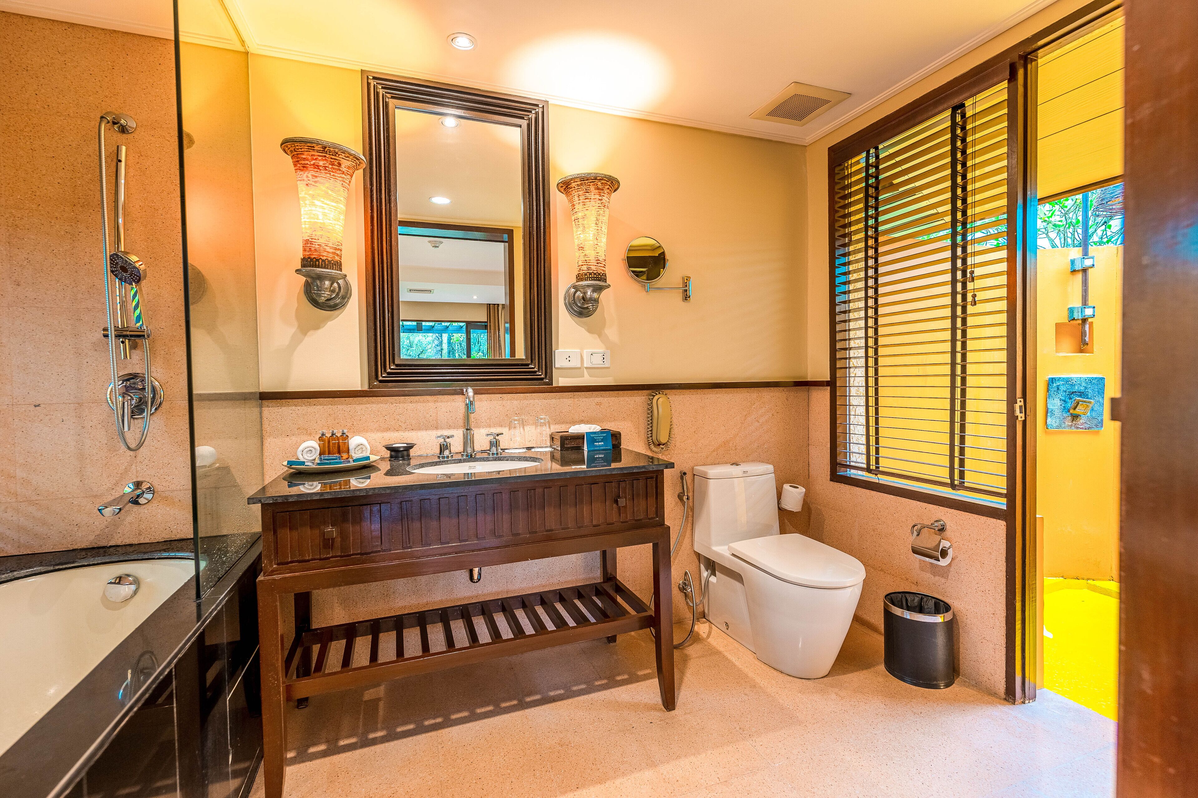 Un baño moderno con bañera de hidromasaje con mampara de vidrio, tocador de madera, inodoro y ventana con persianas.