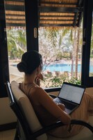 Una persona sentada a una mesa con una computadora portátil, con vista a una piscina rodeada de palmeras.