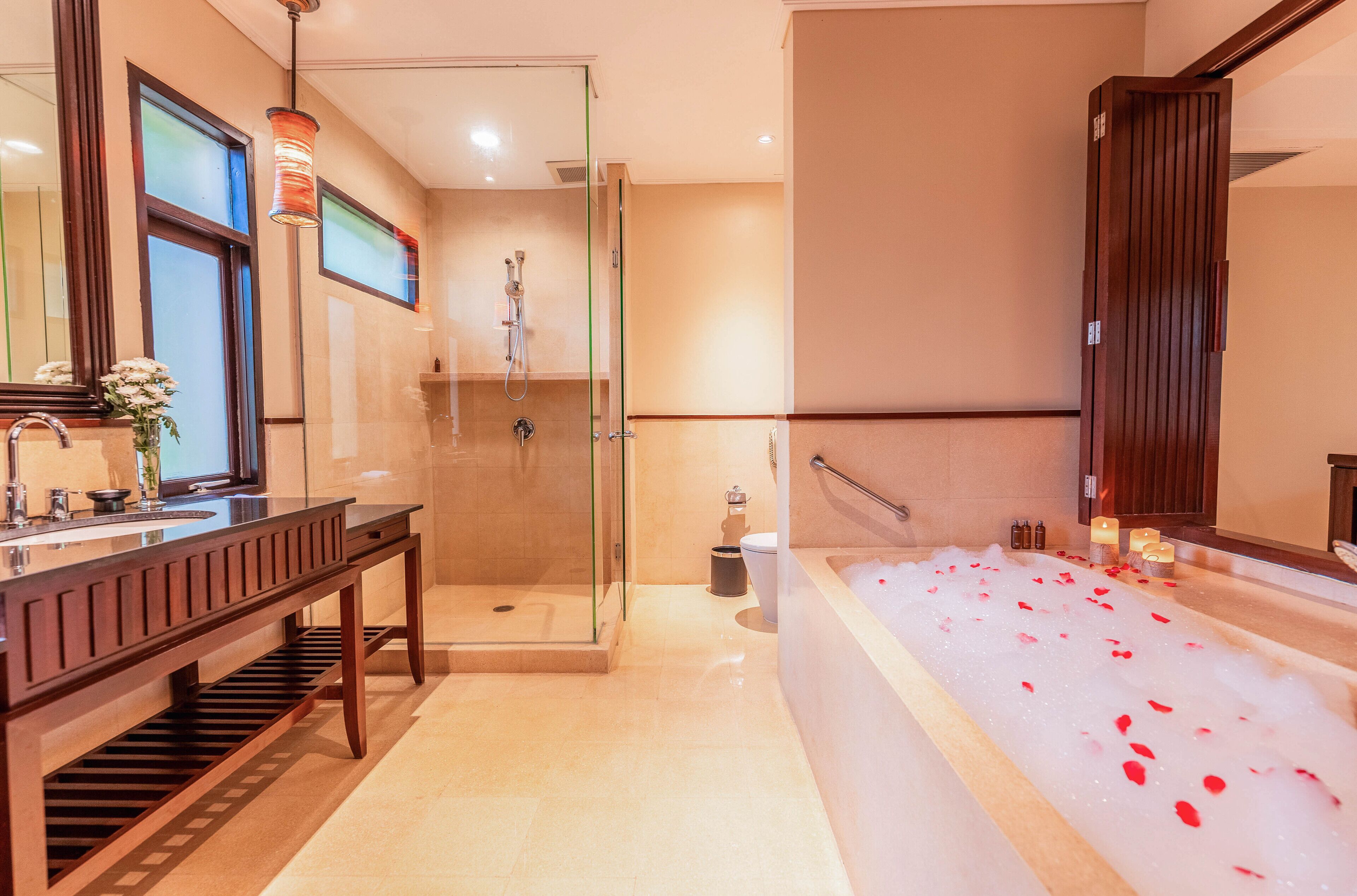 Un bagno moderno con doccia vetrata, un mobile vanitario in legno con una panca, una grande vasca da bagno con petali di rosa e uno specchio con cornice in legno.