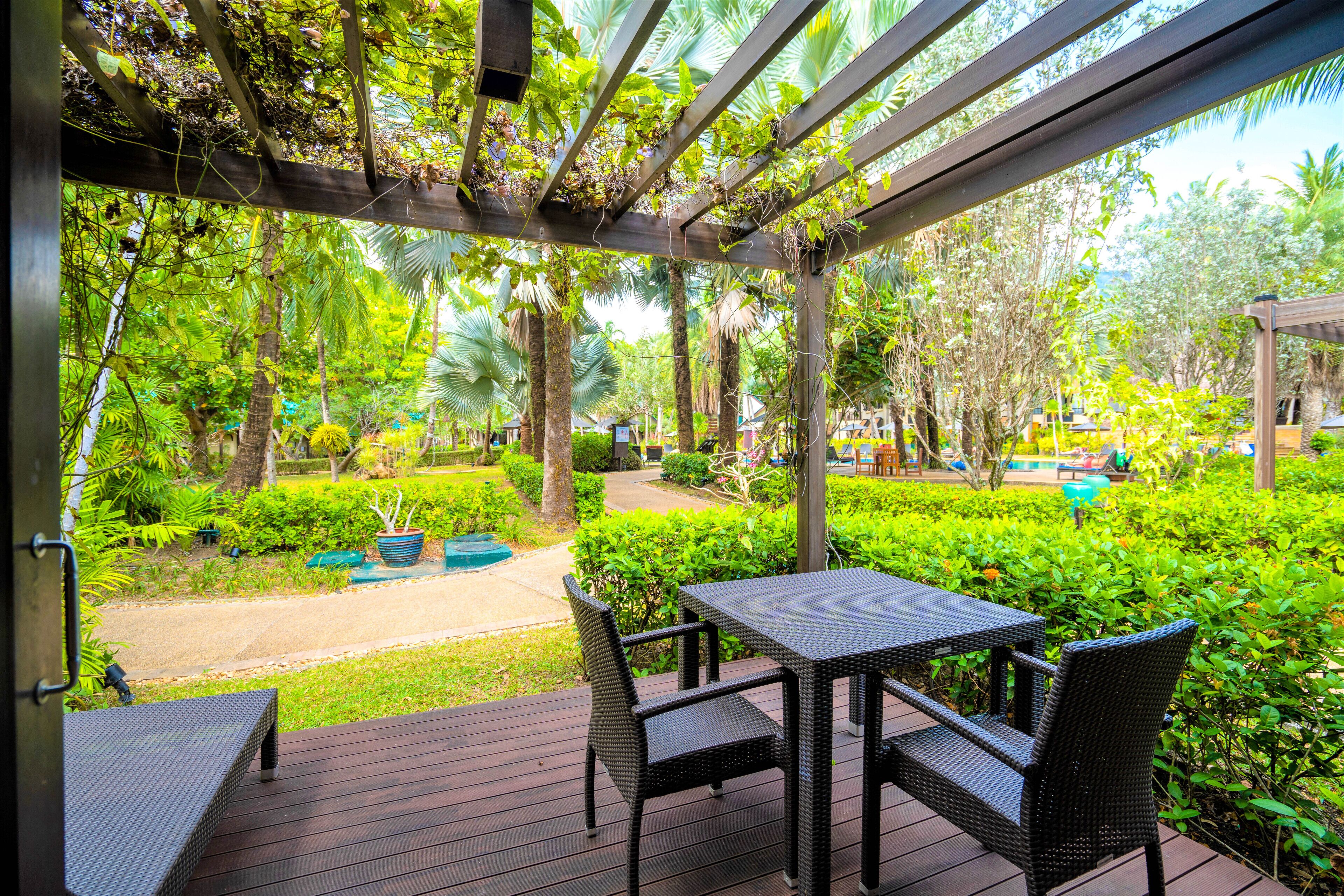 A covered outdoor area with a table and chairs, surrounded by lush greenery and a pool.