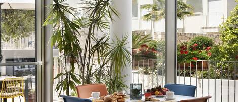 A modern dining area with a table set for four, surrounded by large windows offering a view of a residential building and greenery.